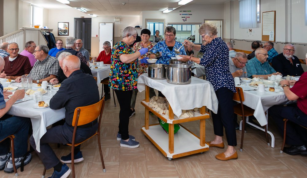 Two women serving food from large silver pots on a serving cart to a large group of men seated at tables in a church hall. The serving women are wearing patterned tops and gloves.