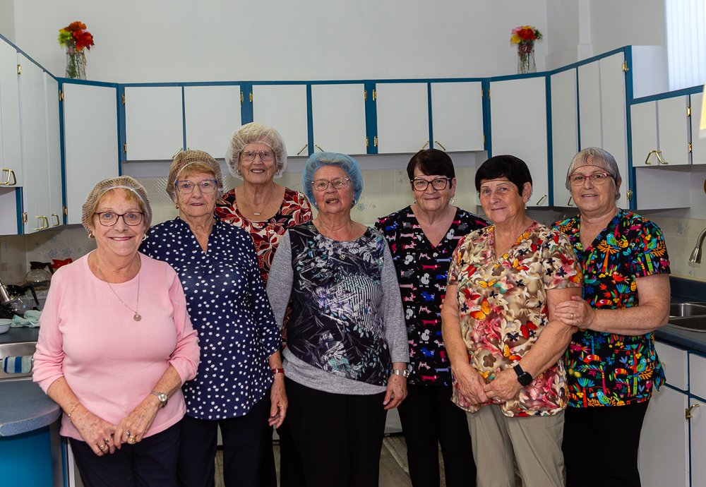 Seven women wearing hairnets and brightly colored tops stand in a row in a church kitchen with white cabinets, posing for a group portrait.