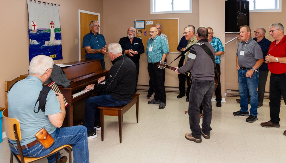 A group of men gathered in a room for a sing-along. One man is playing the upright piano, another is playing a guitar, and several others are standing and singing along with them.