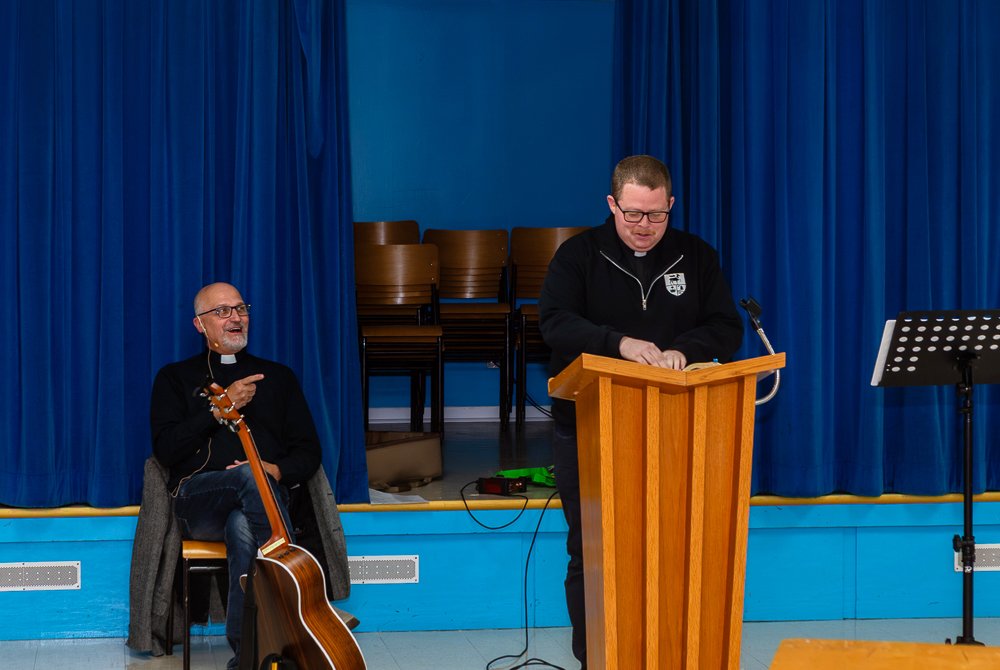 Two men on a stage with a blue curtain backdrop. One man stands at a wooden lectern with an acoustic guitar, and the other man stands to the right singing and playing an acoustic guitar into a microphone with a music stand in front of him.