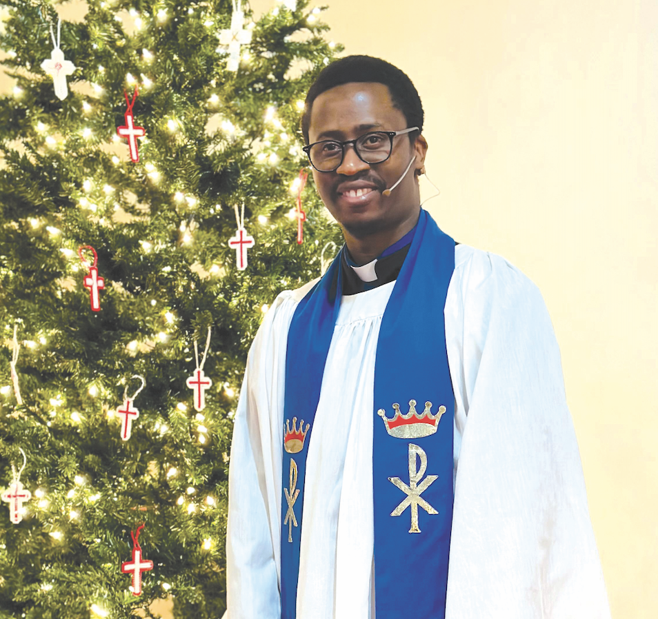 A man dressed as a priest wearing a blue stole, standing in front of a Christmas tree