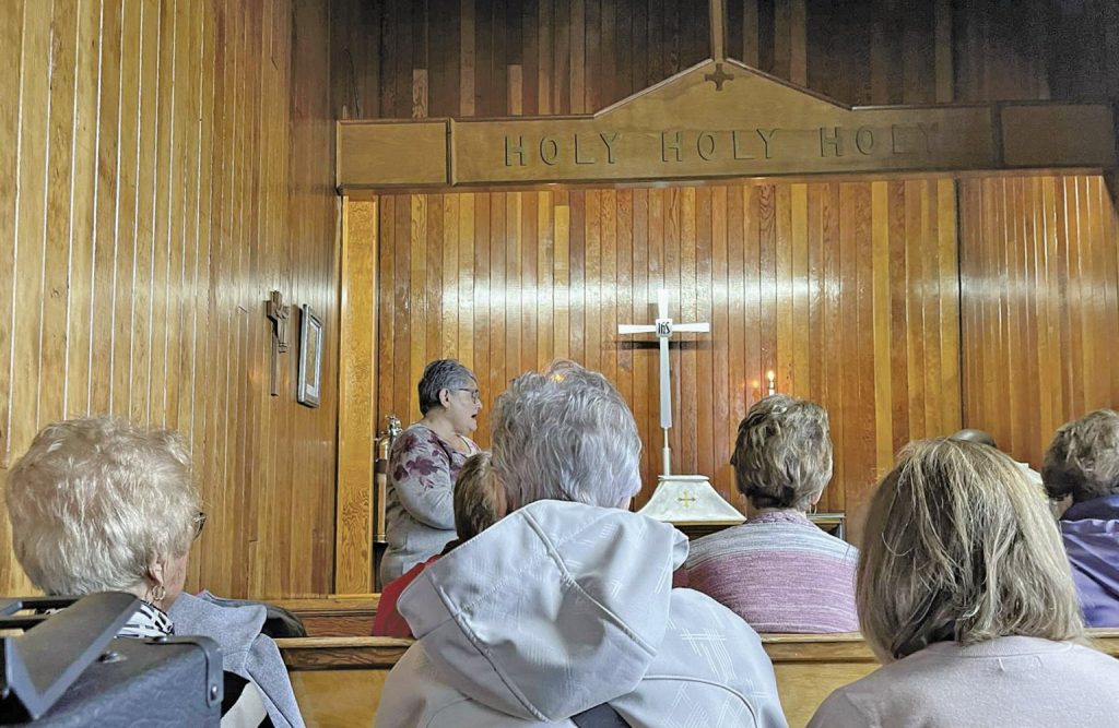 Woman speaking in a church with the word 'HOLY' repeated on the wall.