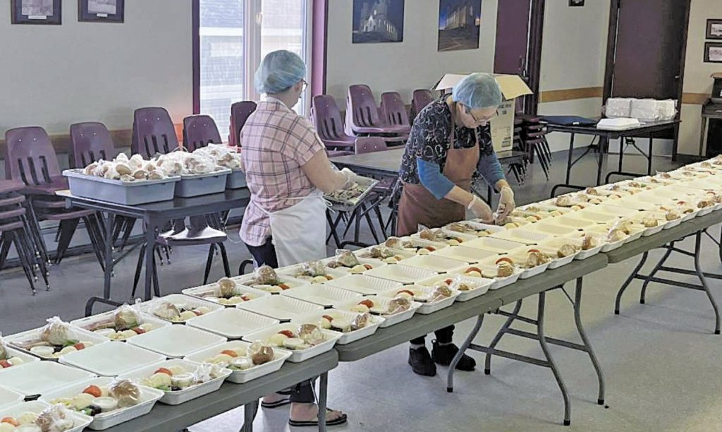 Two women preparing many packaged meals on long tables.