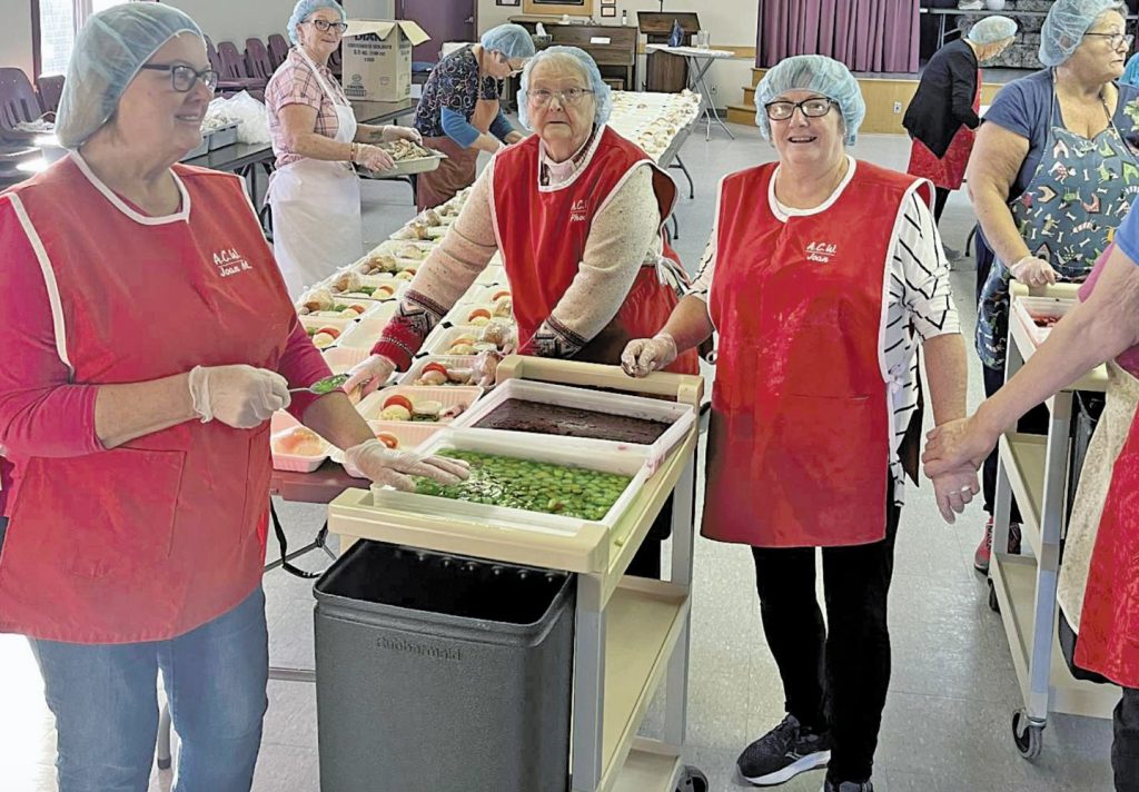 Four women in red aprons and hairnets preparing packaged meals.