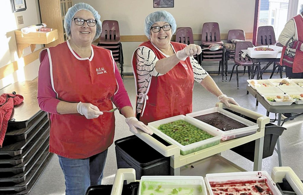 Two smiling women in red aprons and hairnets standing behind a tray of food.