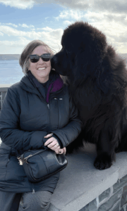 Woman in sunglasses smiling next to a large black dog outdoors.