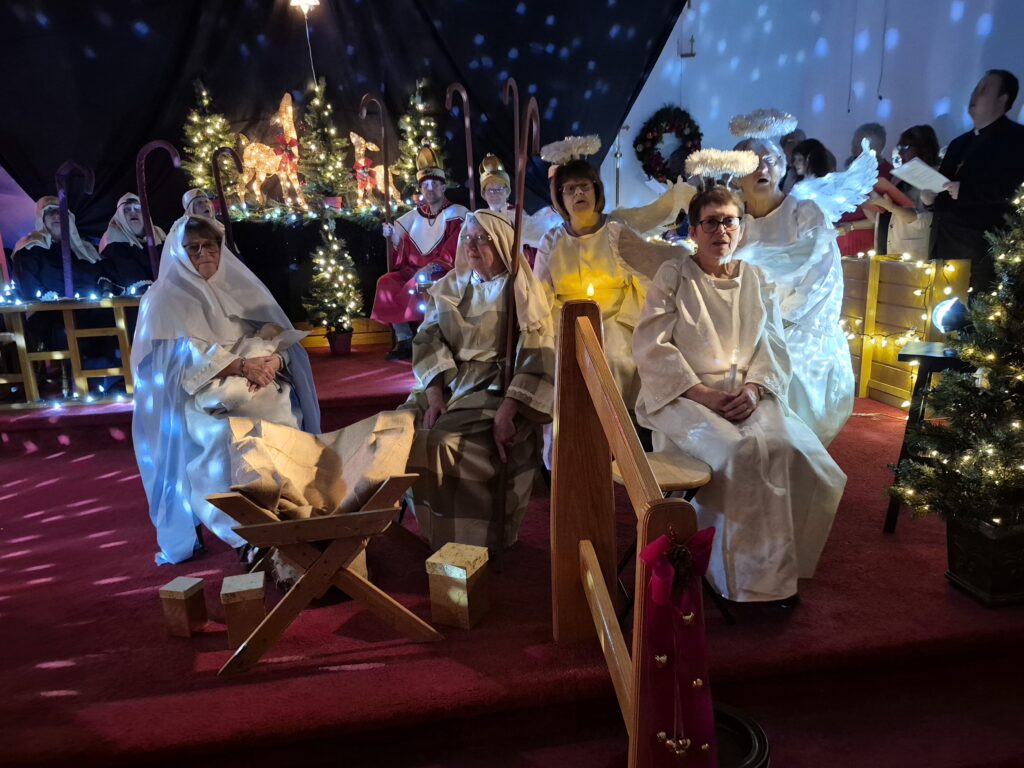 A group of people performing a live nativity play in a church, featuring Mary, Joseph, angels, and shepherds on a decorated stage.