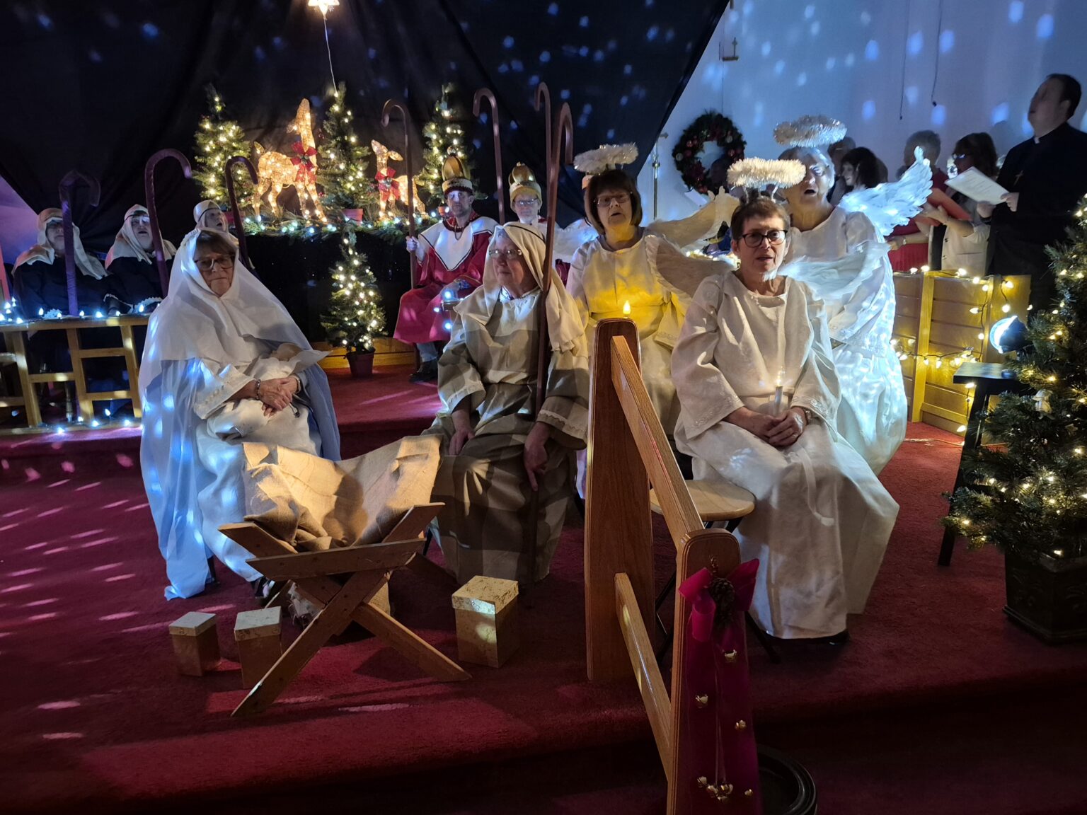 A group of people performing a live nativity play in a church, featuring Mary, Joseph, angels, and shepherds on a decorated stage.
