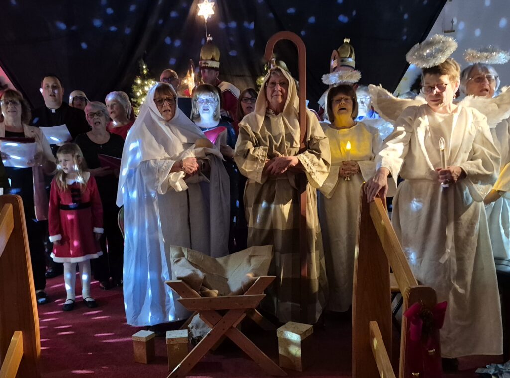 A group of adults in nativity costumes posing behind a wooden manger on a red stage.