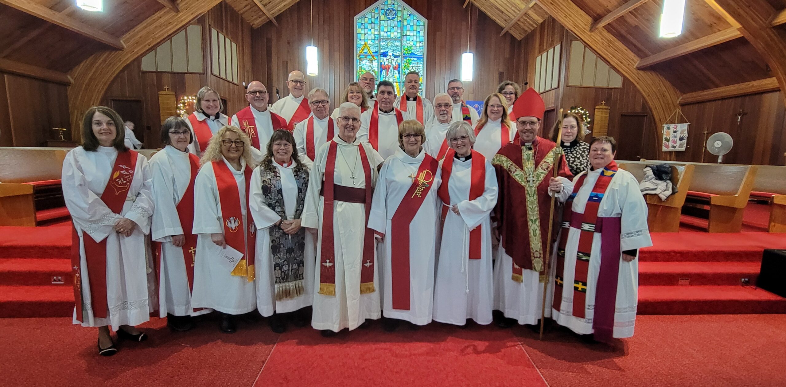 A bishop and a diverse group of clergy members in red and white vestments posing for a group portrait in a wood-paneled church.