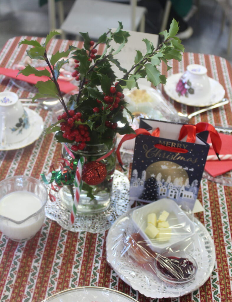 A festive Christmas table setting with a holly centerpiece, teacups, and a small gift bag.