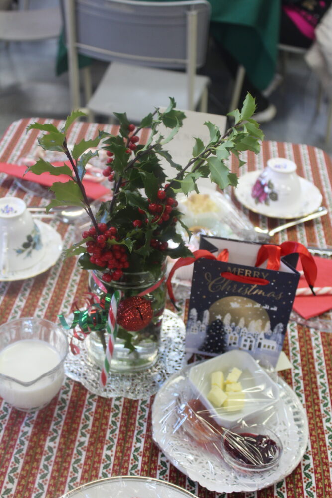 A festive Christmas table setting with a holly centerpiece, teacups, and a small gift bag.