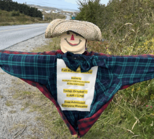 A scarecrow wearing a plaid shirt and straw hat holding a fall festival announcement.