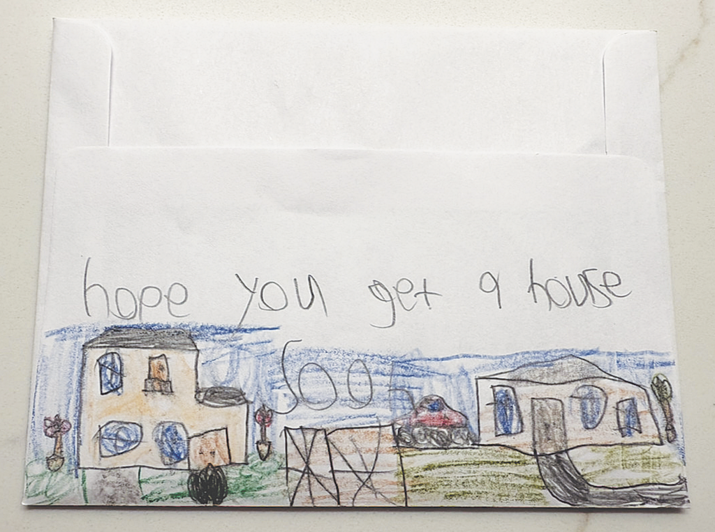 A drawing on an envelope showing two houses and a car with the handwritten message "hope you get a house soon"