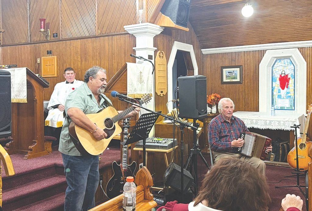 A man playing an acoustic guitar and another man playing an accordion inside a wood-paneled church.