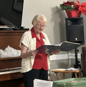 An older woman in a red shirt and white cardigan stands in a living room reading from a large blue binder.