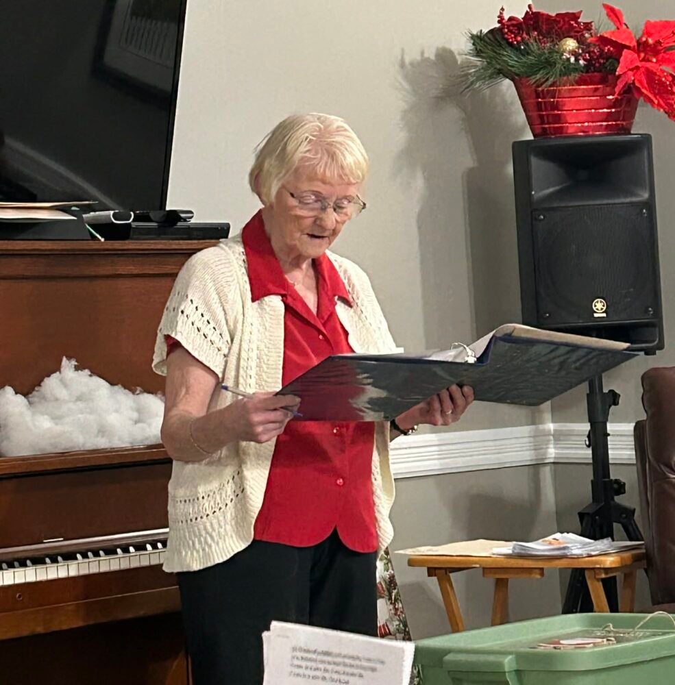 An older woman in a red shirt and white cardigan stands in a living room reading from a large blue binder.