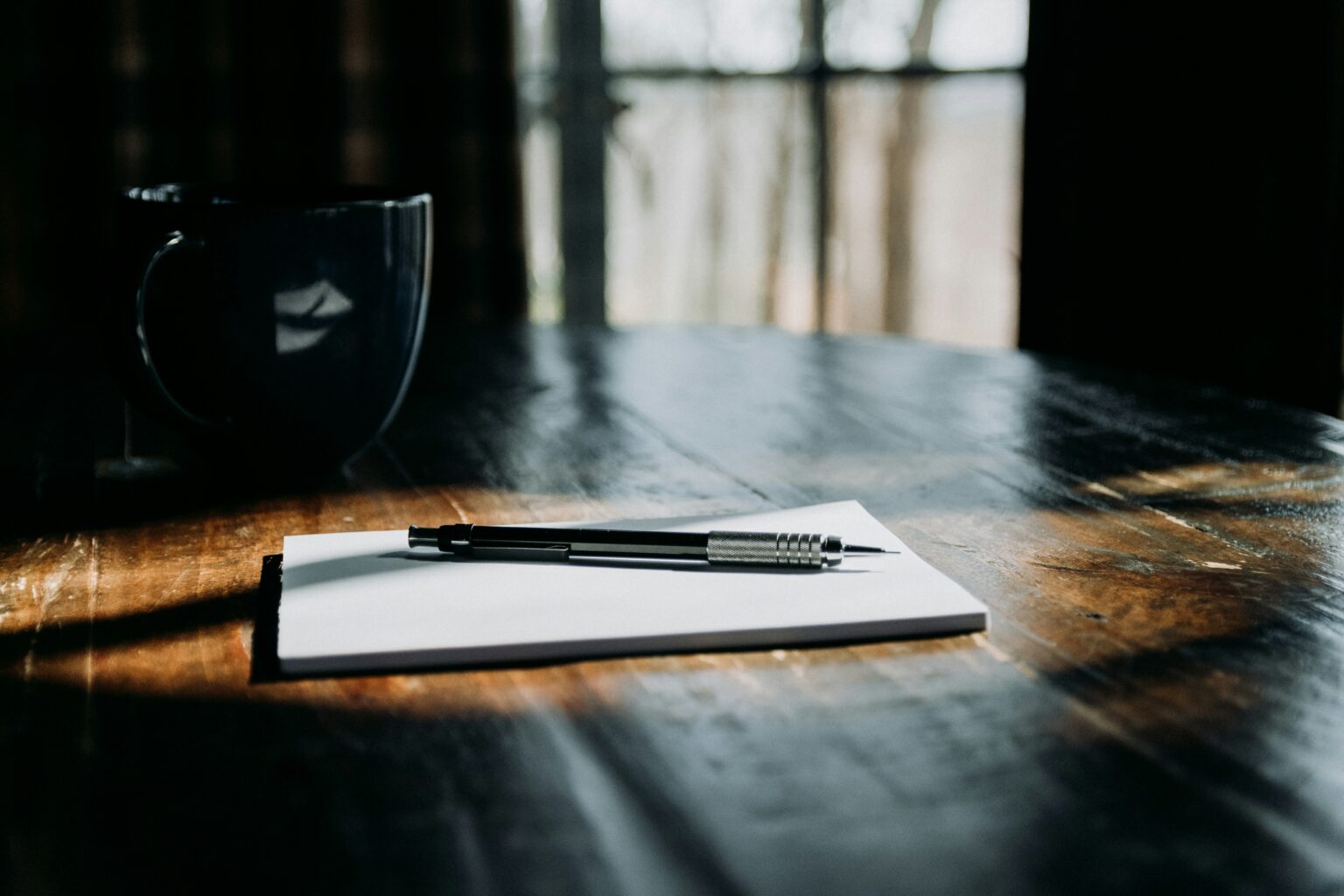 A notebook and mechanical pen on a wooden table, highlighted by natural sunlight next to a dark mug.