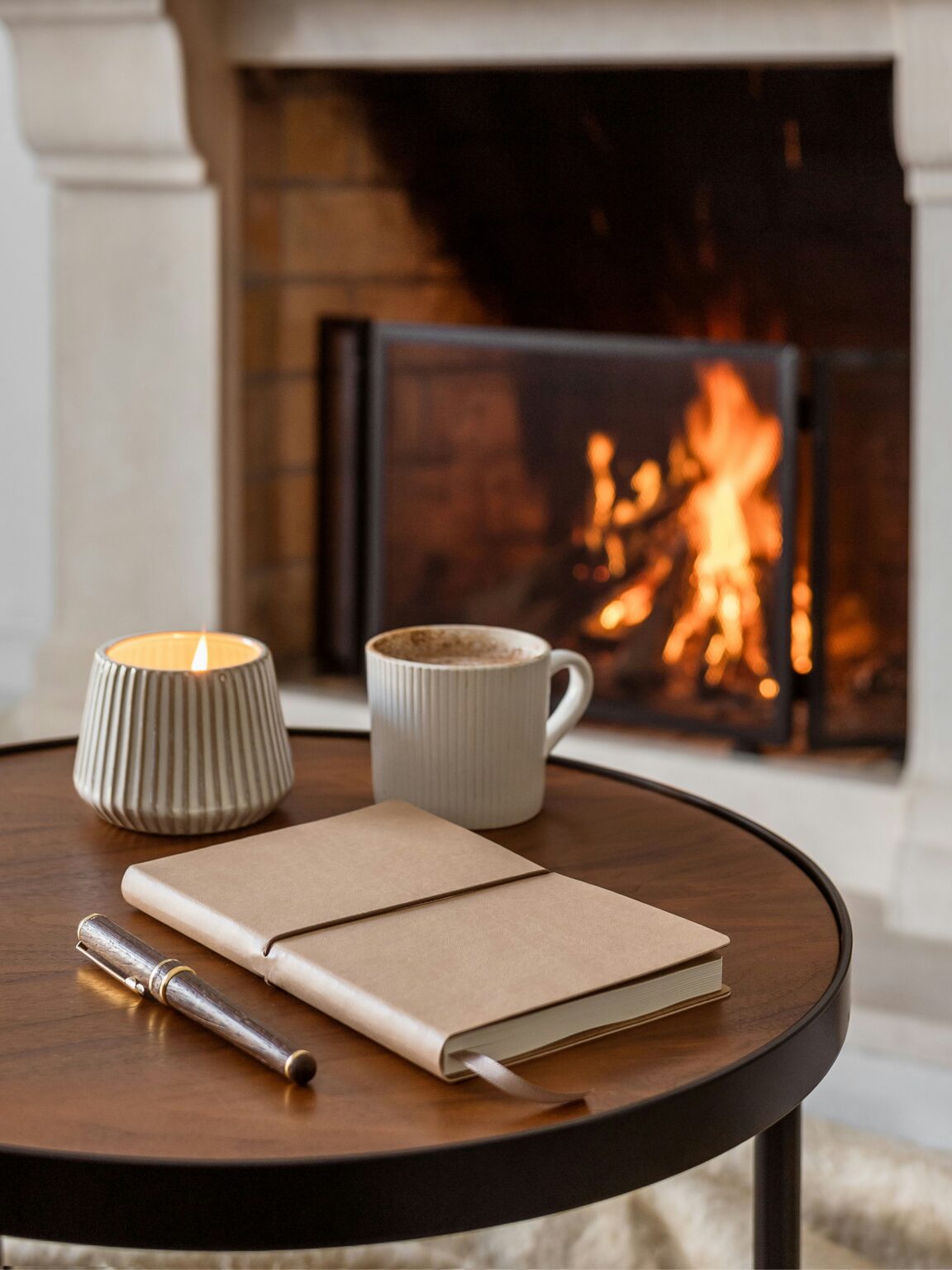A notebook, pen, mug, and lit candle on a table in front of a cozy fireplace.