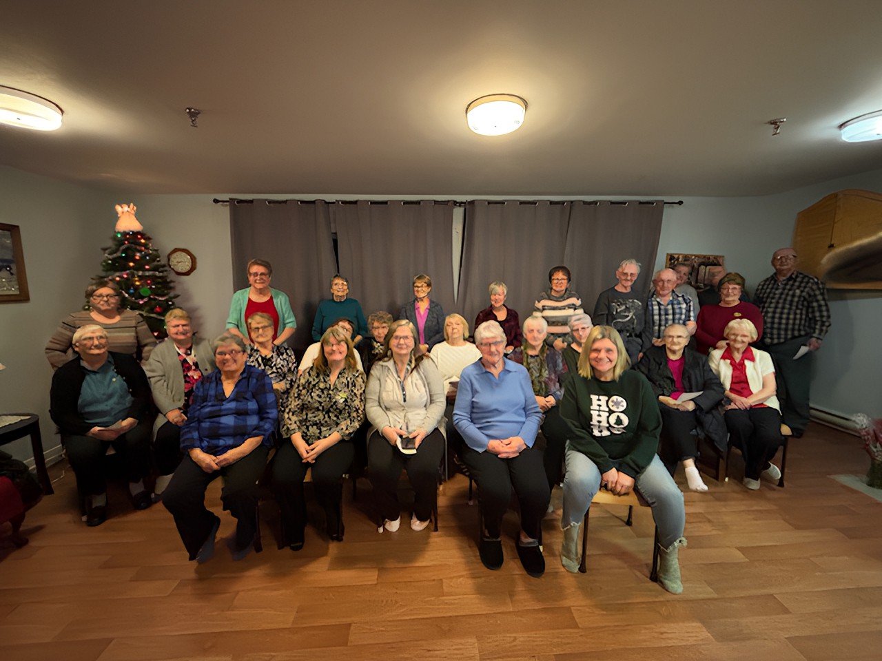 A large group of seniors poses for a photo in a living room decorated with a lit Christmas tree.