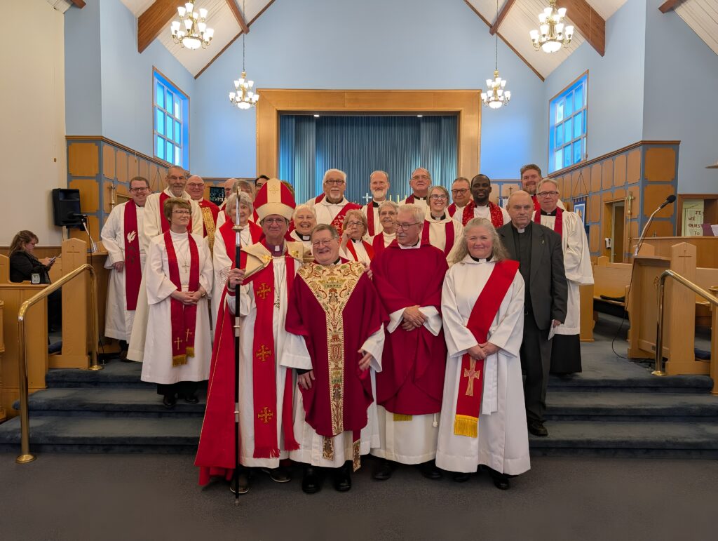 A bishop and a large group of clergy in various vestments pose for a group portrait in a modern church.