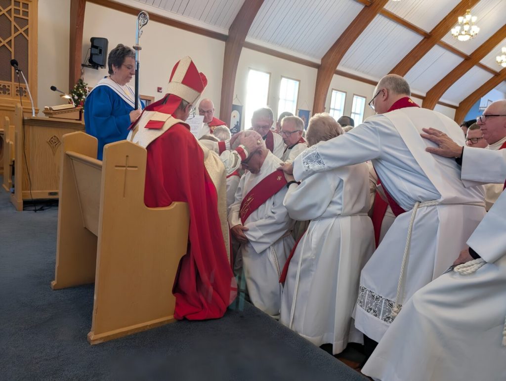 A bishop and several clergy members gather around a kneeling man for a prayer of ordination.