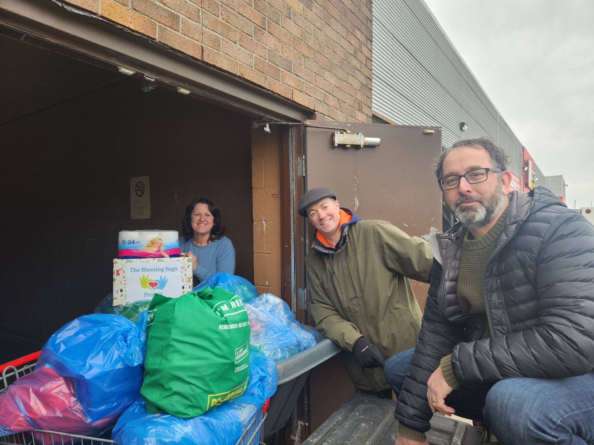 Three people smiling while unloading bags of supplies from a truck into a building.