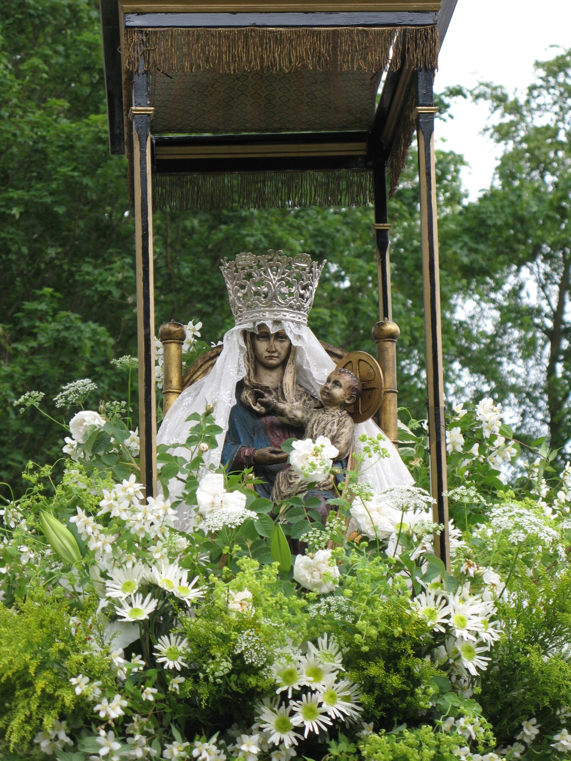 A statue of the Virgin Mary holding the infant Jesus, adorned with a silver crown and white veil, surrounded by white flowers under a fringed canopy.