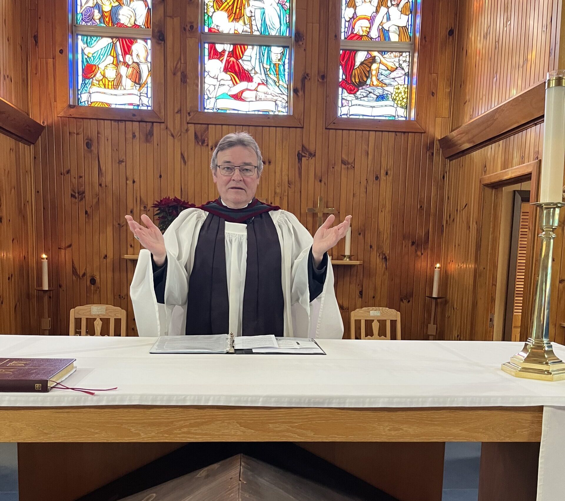 A priest in vestments stands behind a white-covered altar with arms outstretched in a wood-paneled church with stained glass windows.
