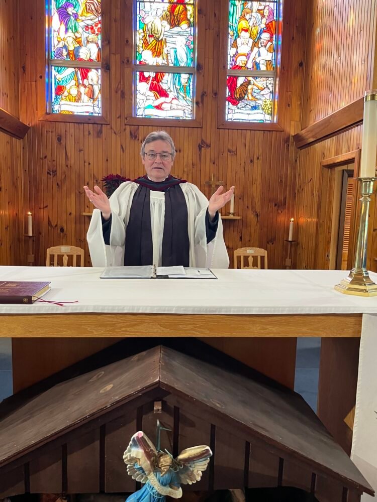 A priest in vestments stands behind a white-covered altar with arms outstretched in a wood-paneled church with stained glass windows.