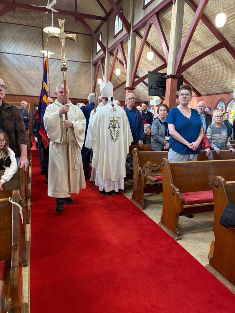 A church procession featuring a person carrying a tall golden cross down a red-carpeted aisle between wooden pews.