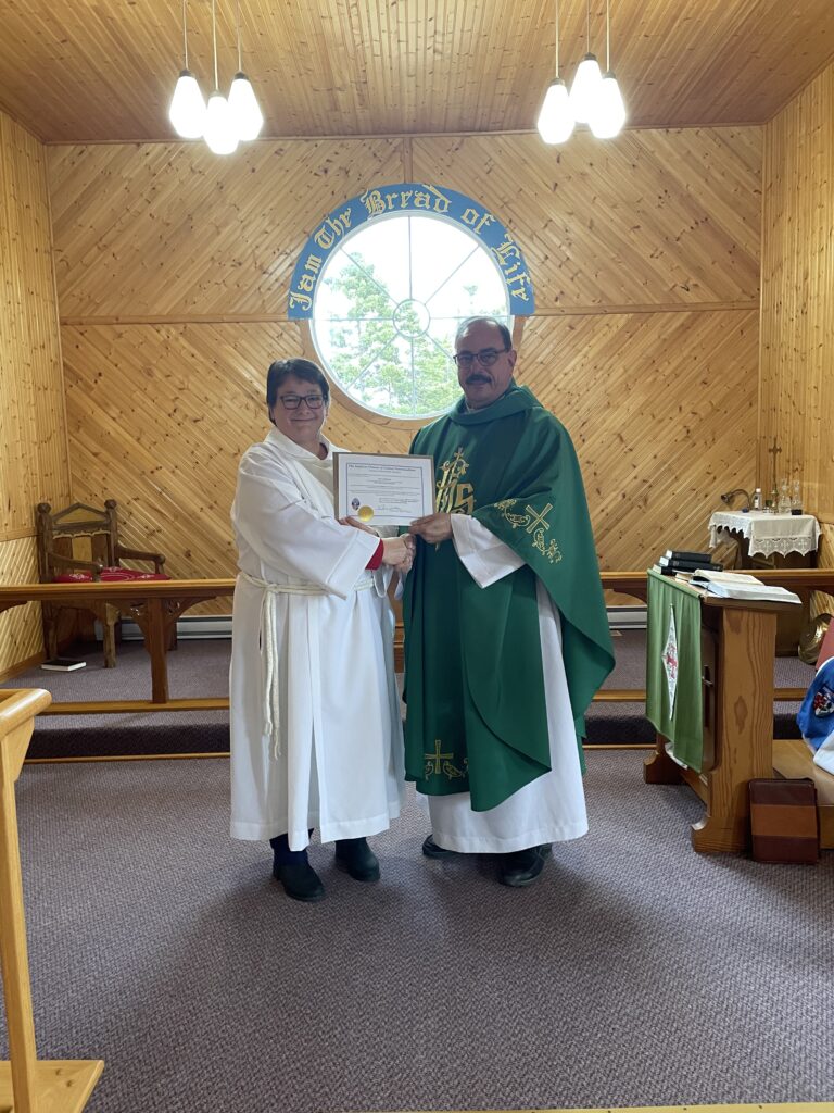 A priest in a green chasuble and a woman in a white alb stand together in a wood-paneled church sanctuary holding a certificate.