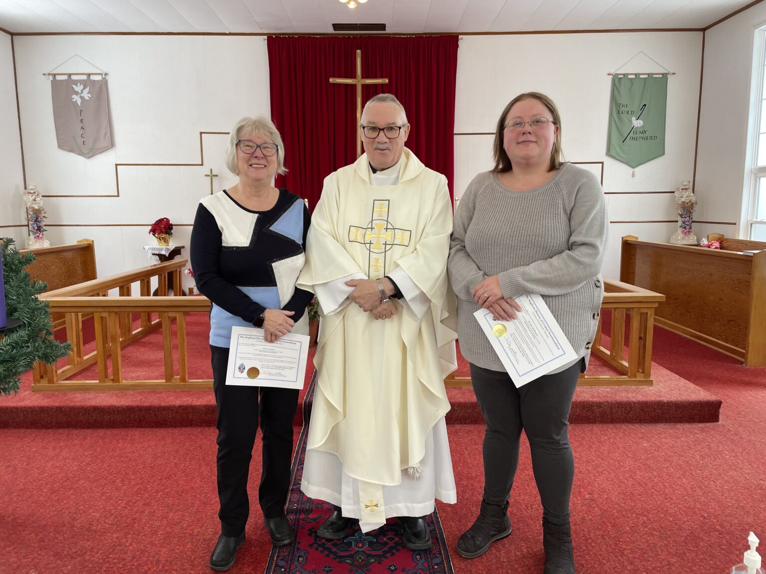 A priest in white vestments stands between two women holding certificates in a church with red carpeting.