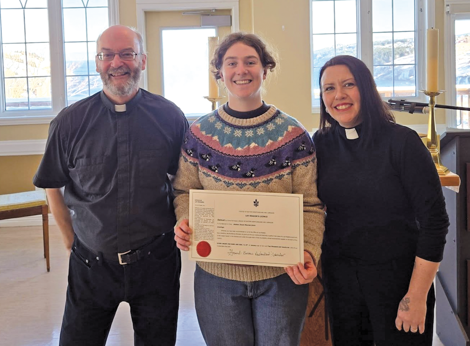 Three smiling people standing indoors, with the person in the center holding an official "Lay Reader's Licence" certificate.