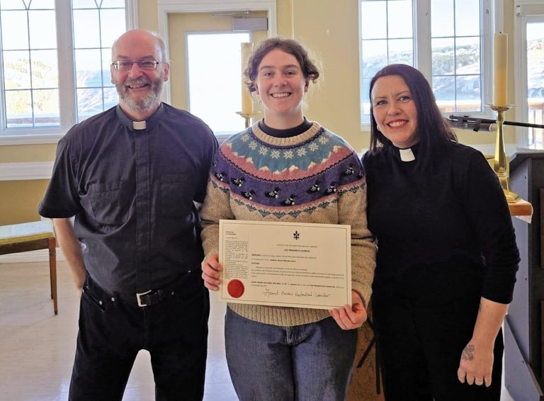 Three smiling people standing indoors, with the person in the center holding an official "Lay Reader's Licence" certificate.