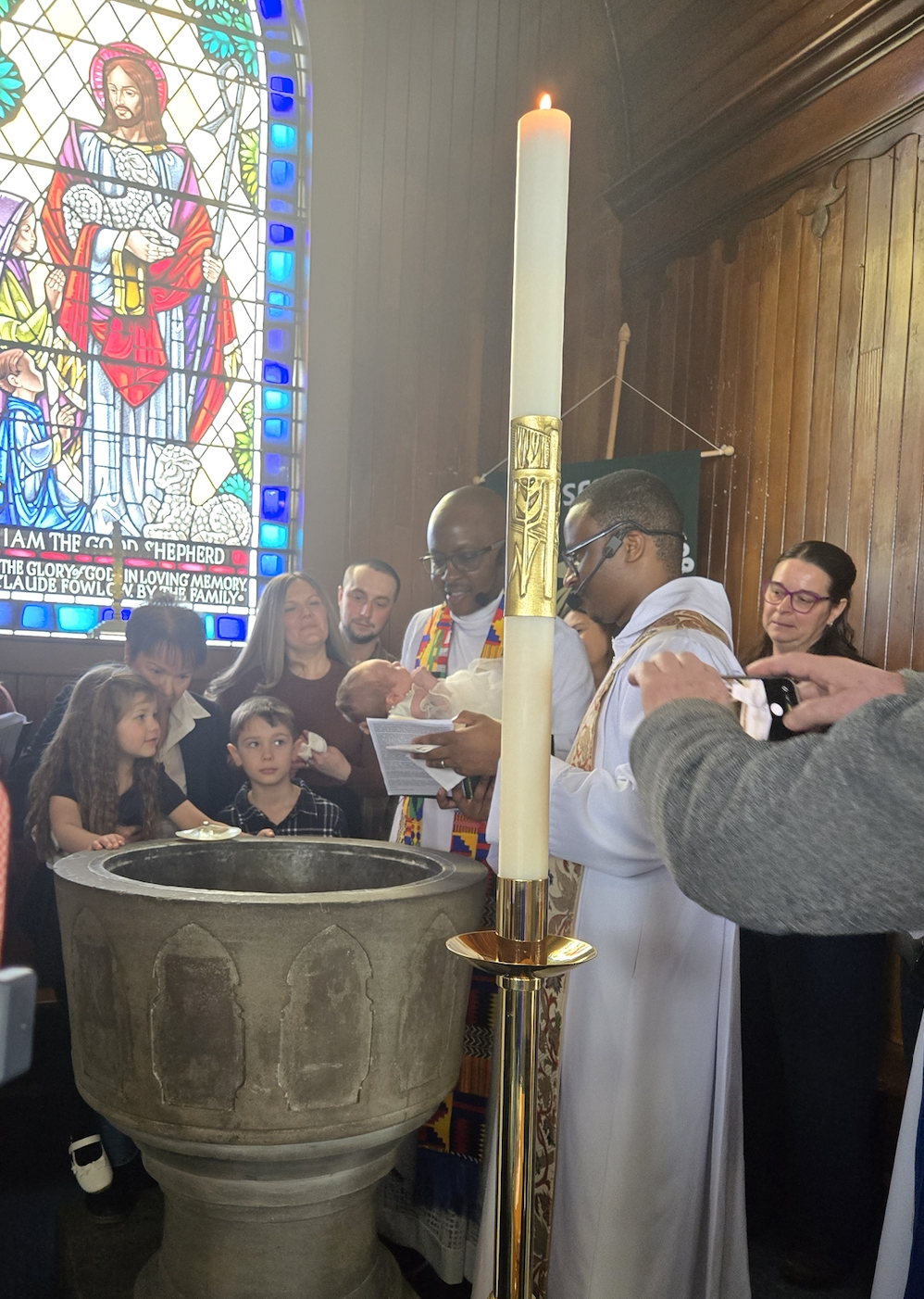 A group of people gather around a stone baptismal font in a church during a ceremony.