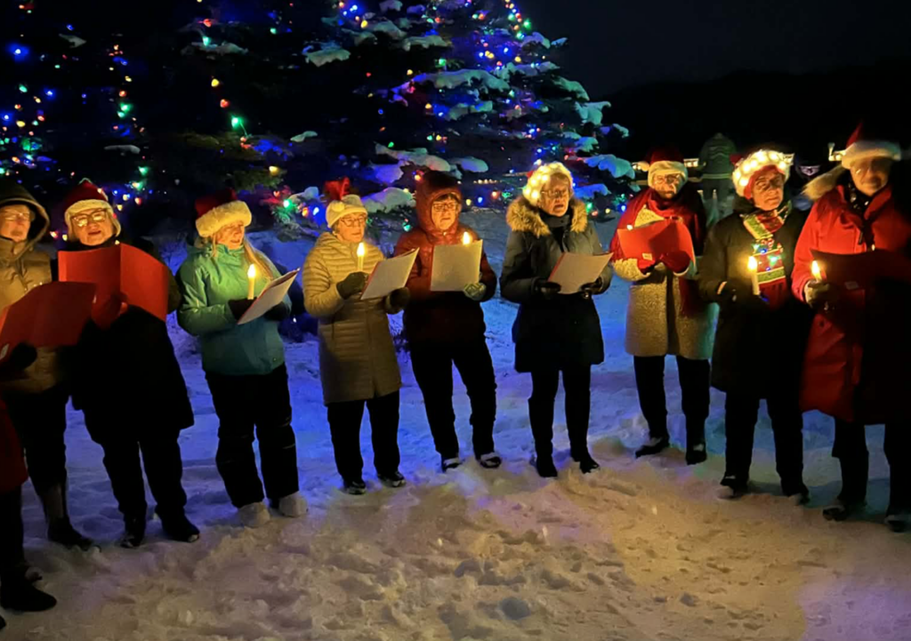 a group of people singing in front of a lit Christmas tree, outside in the snow