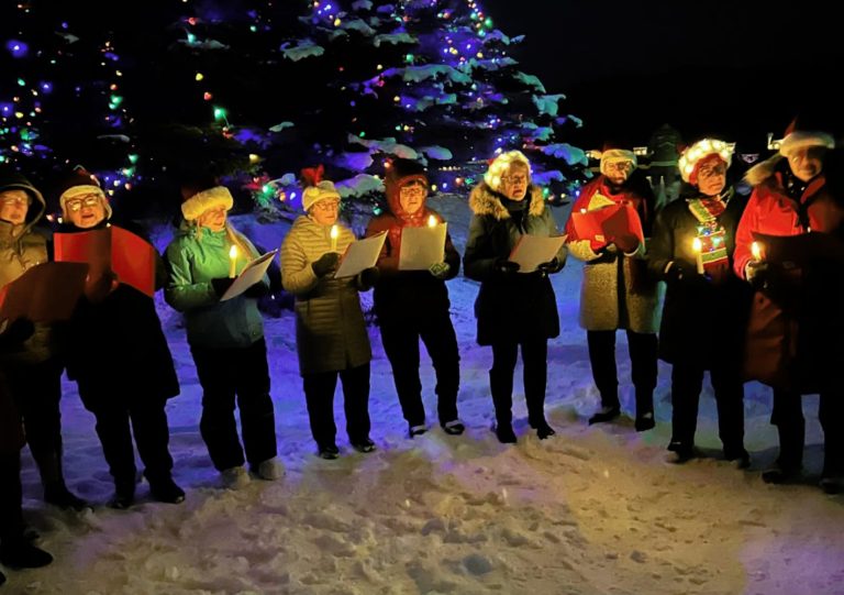 a group of people singing in front of a lit Christmas tree, outside in the snow