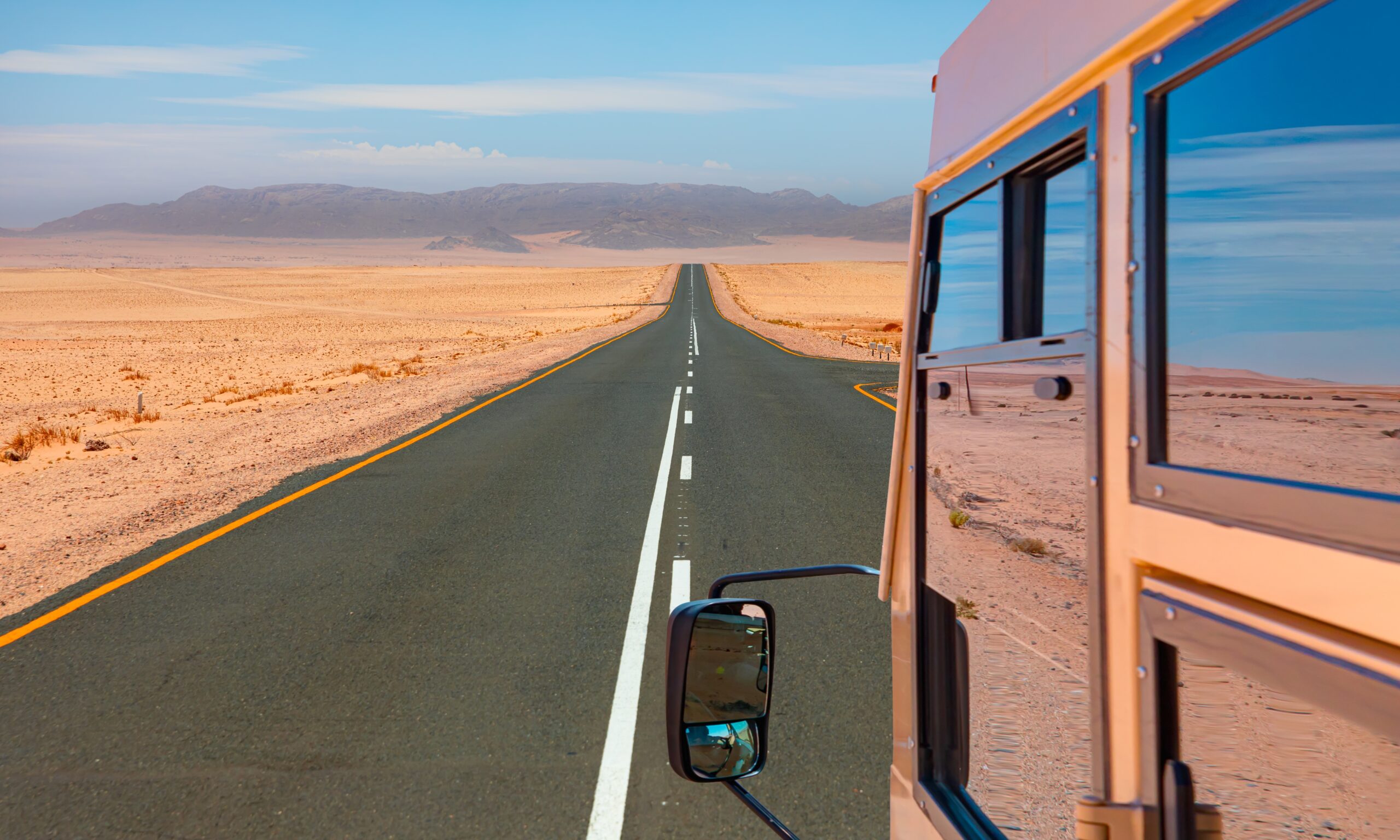 A long, straight two-lane highway stretching through a vast, arid desert landscape as seen from the side of a bus.