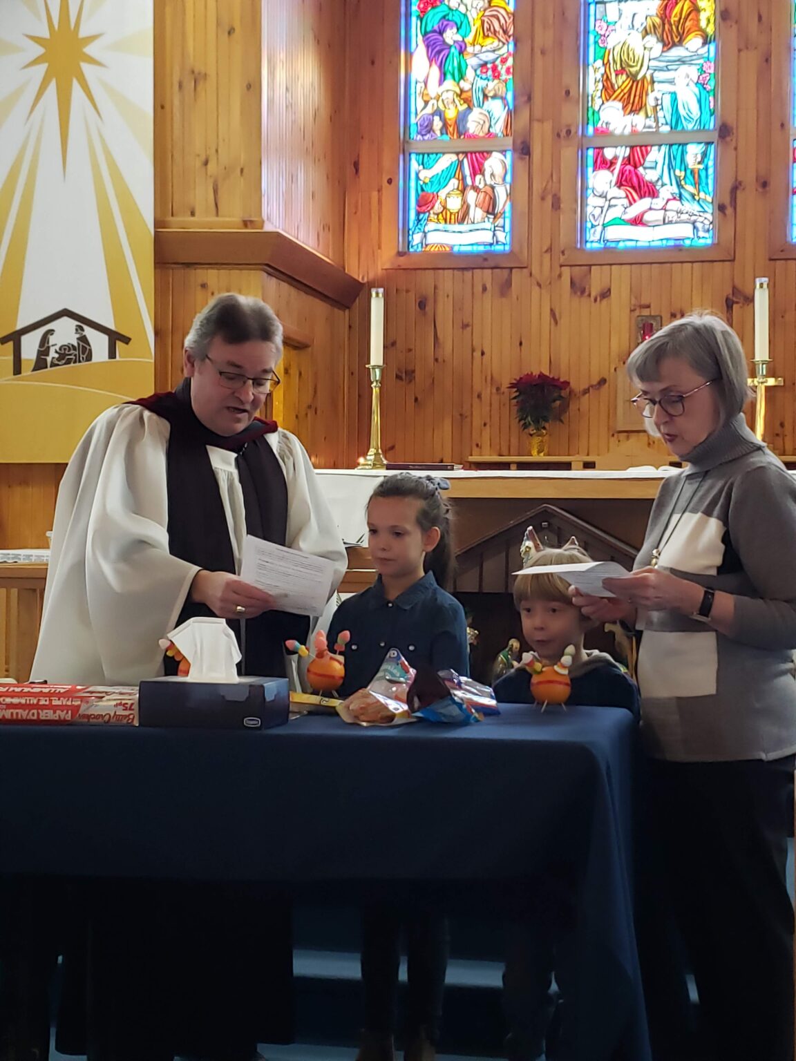 a man in clerical vestments shows two children and a woman oranges on a table at the front of a wooden church