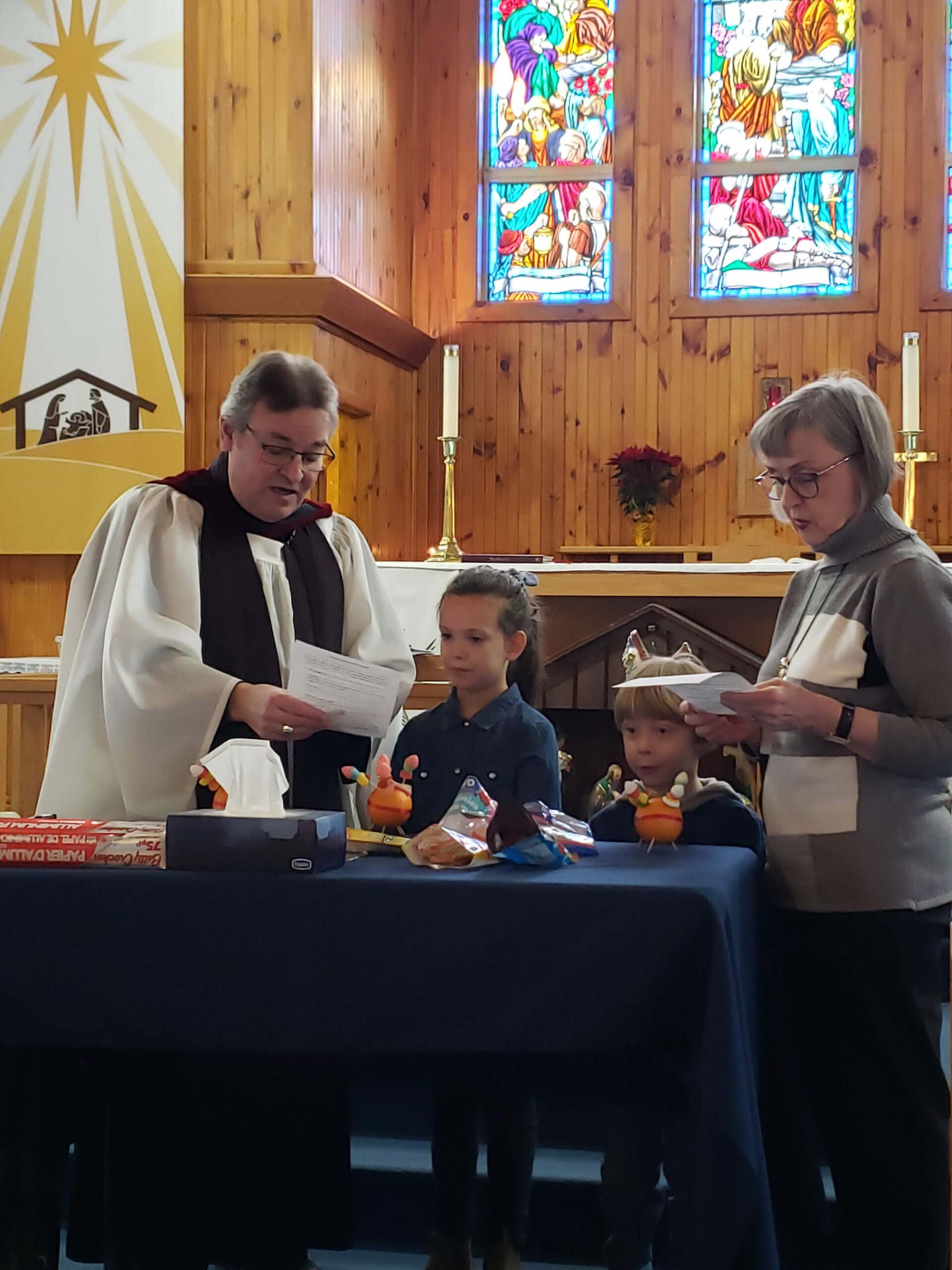 a man in clerical vestments shows two children and a woman oranges on a table at the front of a wooden church