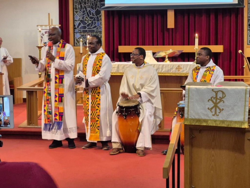 A group of clergy, dressed in white vestments and colourful stoles, some playing traditional African drums, at the front of a church