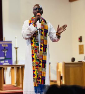 a man preaches a sermon at the front of a church dressed in white vestments and a colourful stole