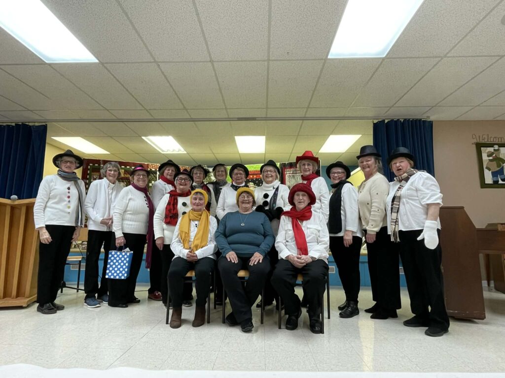 A group portrait of seventeen women dressed in coordinated white and black outfits with colorful winter accessories, posing in front of a stage.