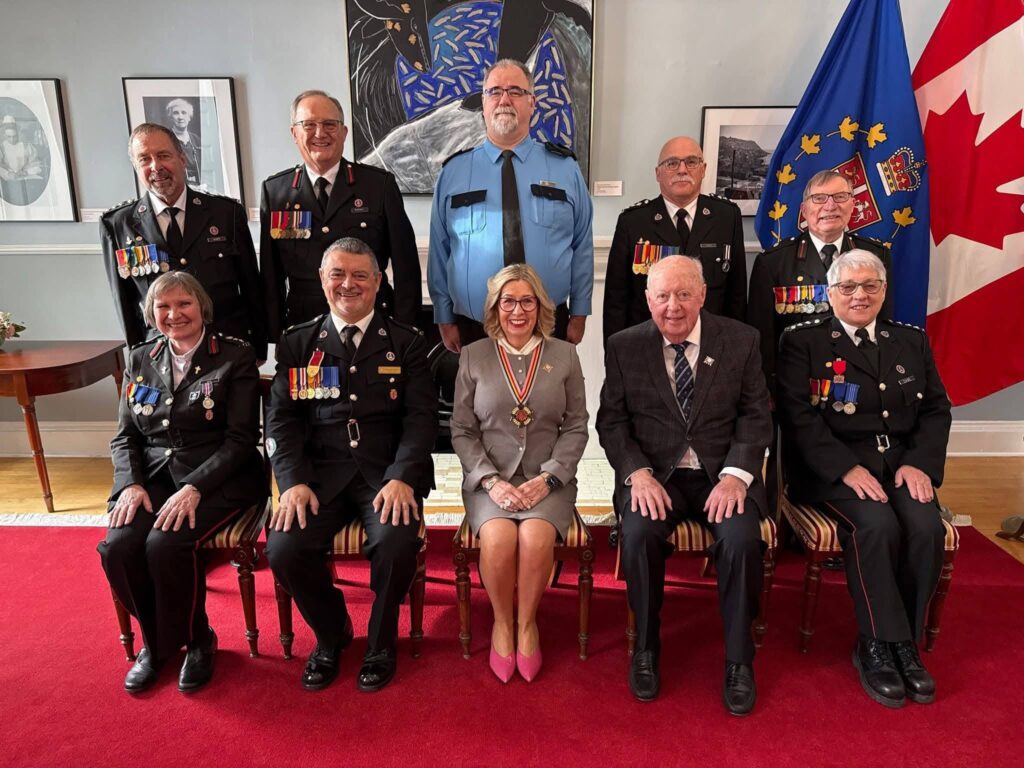 A group of nine people, many in dress uniforms with medals, posed for a formal photo in a room with a red carpet and a Canadian flag.