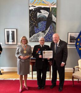 A person in a black dress uniform holding an award certificate, standing between a woman in a grey suit and a man in a plaid jacket.