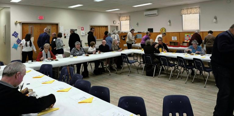 A room filled with people socializing, getting food from a buffet, and seated at long tables.