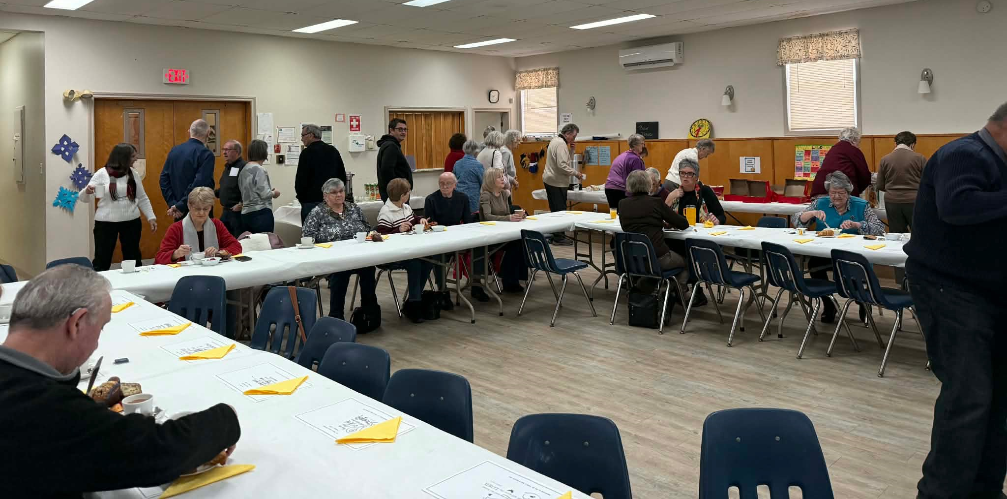 A room filled with people socializing, getting food from a buffet, and seated at long tables.