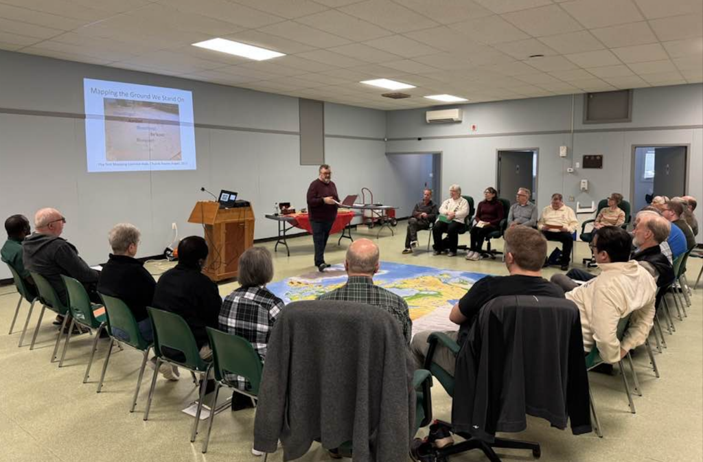 A man leads a presentation titled "Mapping the Ground We Stand On" for a group seated in a large circle around a floor map.