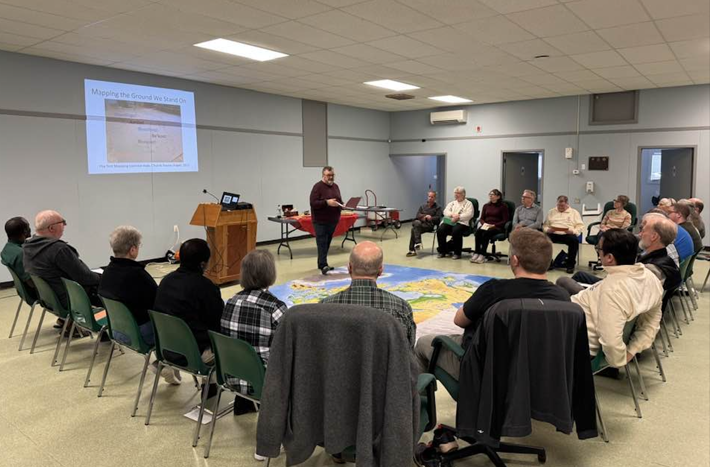 A man leads a presentation titled "Mapping the Ground We Stand On" for a group seated in a large circle around a floor map.